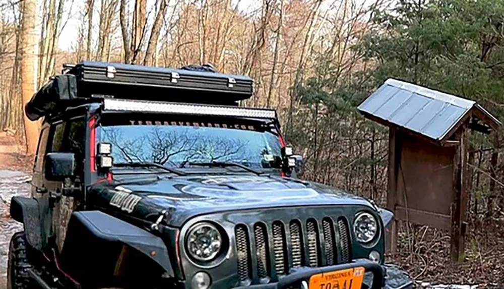 A rugged off-road vehicle equipped with roof-mounted gear is parked in a woodland area near a simple wooden structure with a solar panel on its roof