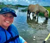 A smiling person in a life jacket takes a selfie while kayaking in a grassy waterway with a horse wading and grazing in the background