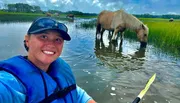 A smiling person in a life jacket takes a selfie while kayaking in a grassy waterway with a horse wading and grazing in the background.