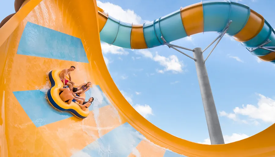 Four individuals are enjoying a thrilling ride down a bright yellow water slide in a blue raft.