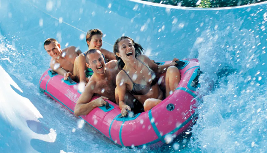 Four people are enjoying a thrilling ride down a water slide in a pink inflatable raft, splashing water around and laughing.