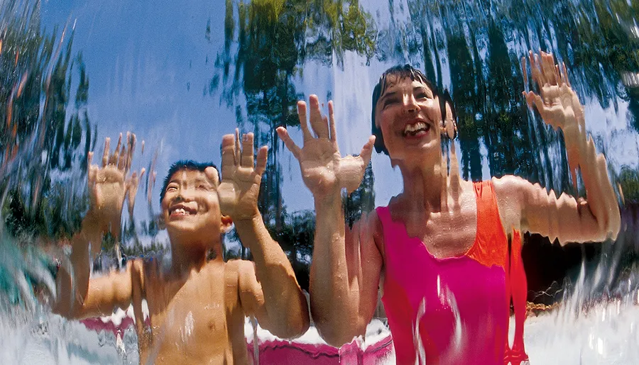 A young boy and a woman are smiling and reaching out towards the camera through a clear curtain of water, likely enjoying a sunny day at a water play area.