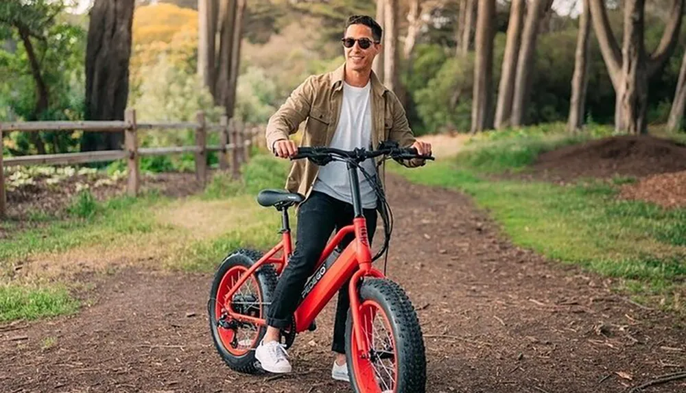 A person is smiling while standing over a red electric bicycle on a dirt path in a wooded area