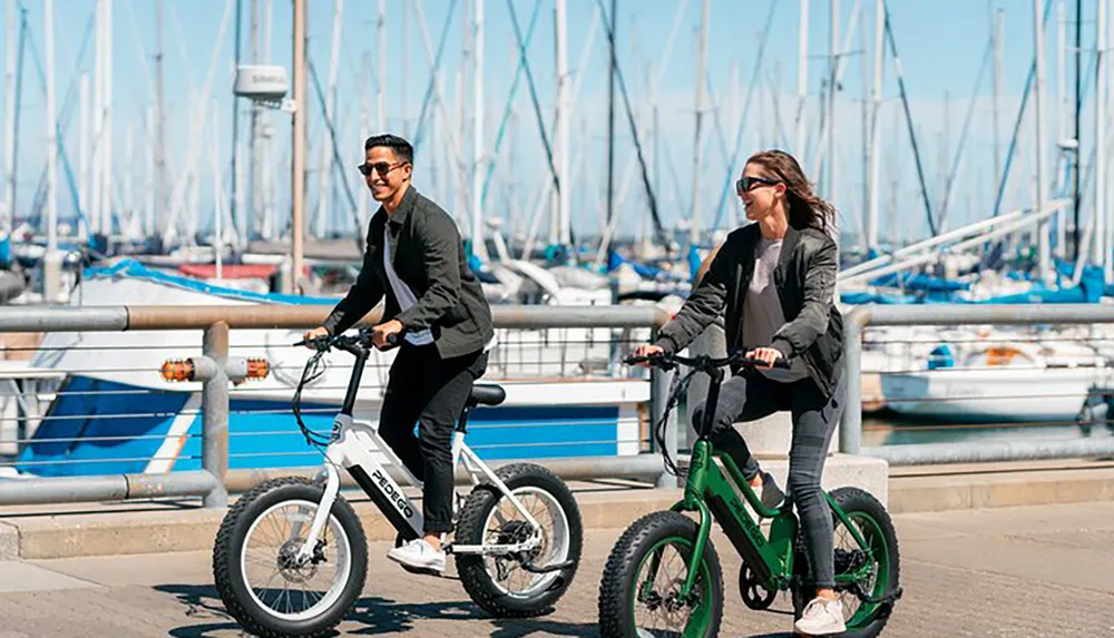 Two people are riding electric bikes along a harbor filled with docked sailboats on a sunny day