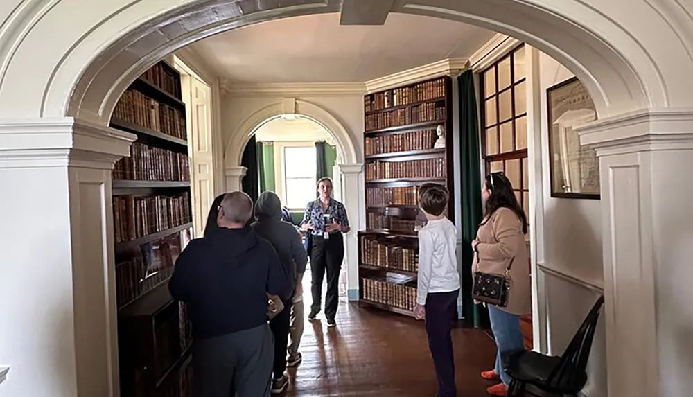 A group of people is on a tour in an elegant room with arched doorways and wall-to-ceiling bookshelves filled with old books