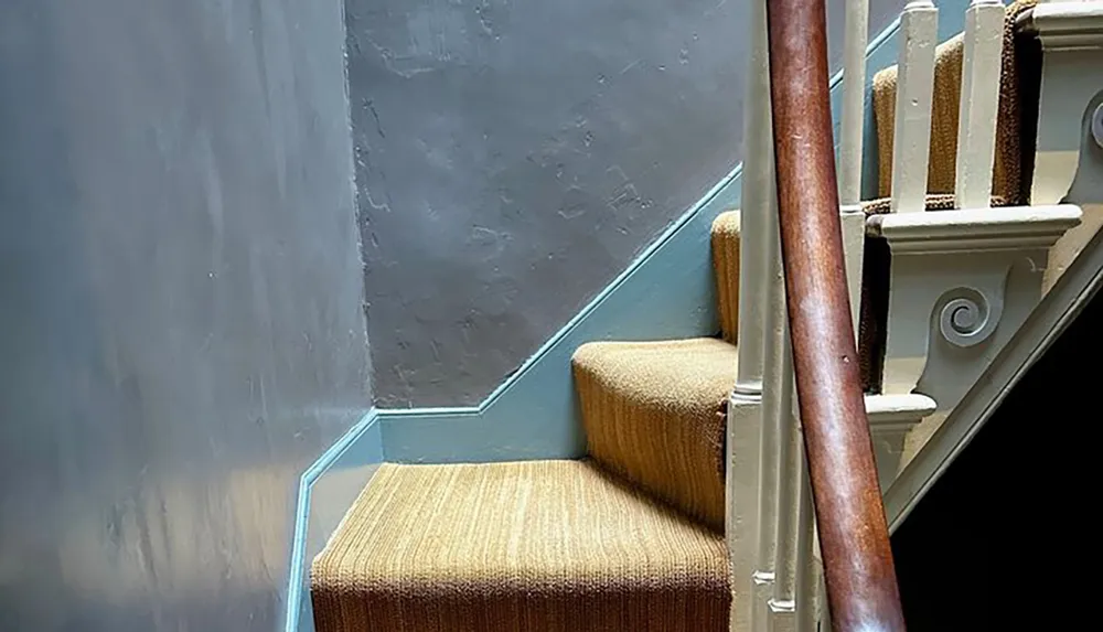 A carpeted staircase with a wooden handrail and white balusters is illuminated by a light source creating a blue trim illusion along its edge