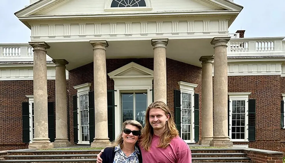 A woman and a man are smiling in front of a classical brick building with grand columns and a pediment