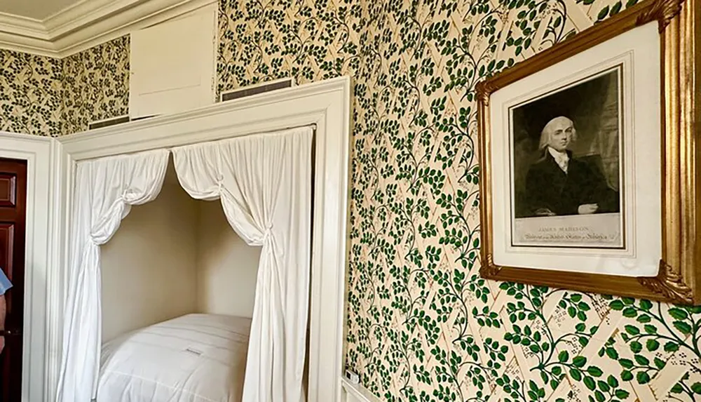 A historical room with a green leaf-patterned wallpaper features a built-in alcove bed with white drapery and a framed portrait of a distinguished individual on the wall