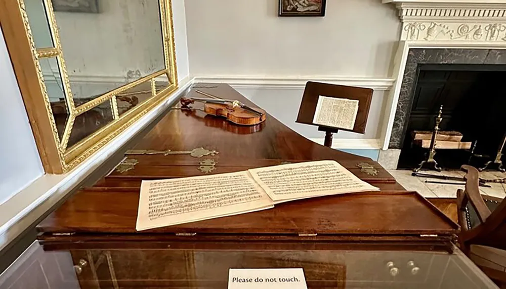 The image shows a historical music room with a violin on a desk sheet music a music stand a fireplace and a sign that reads Please do not touch