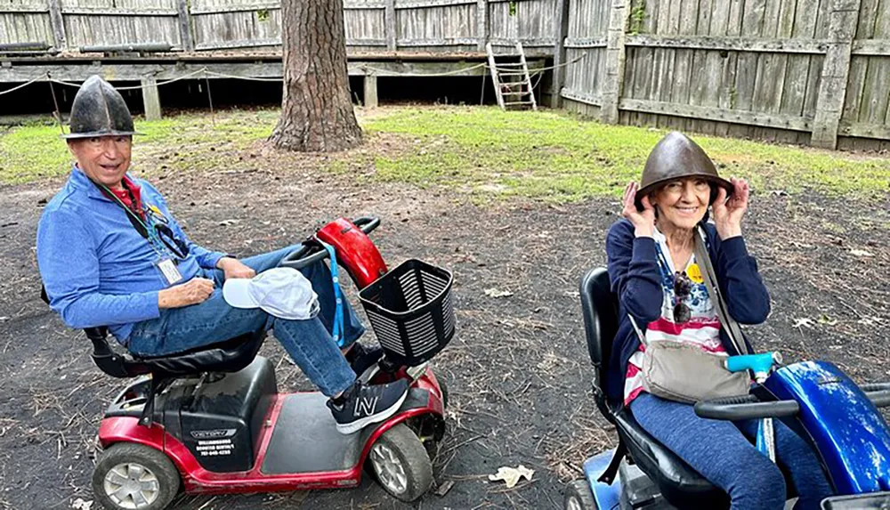 Two smiling individuals are wearing old-fashioned metal helmets while seated on mobility scooters outdoors