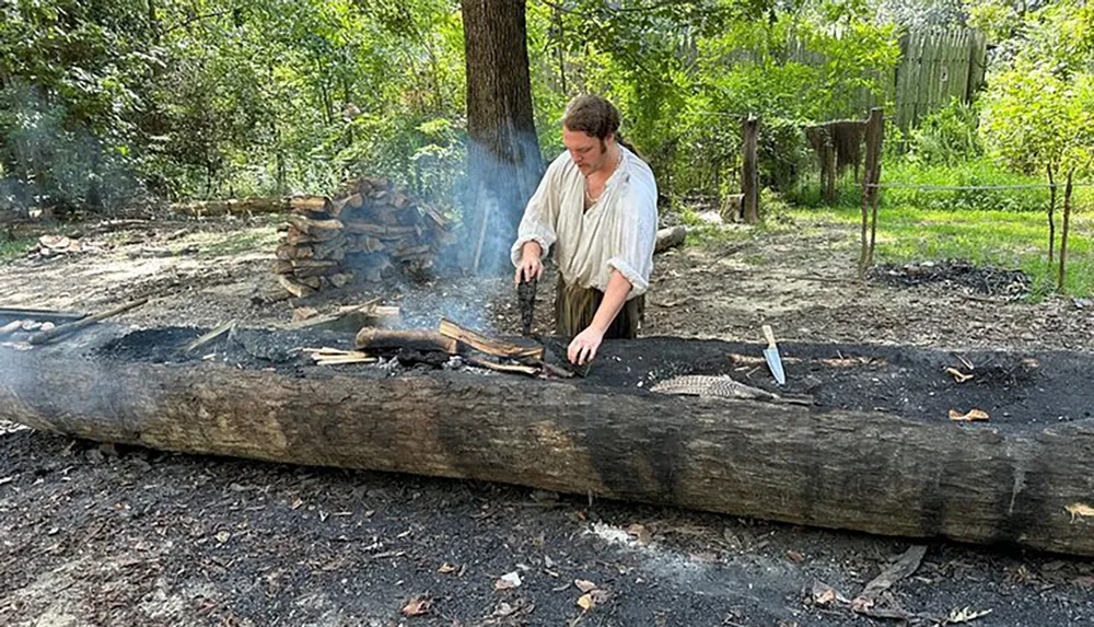 A person in historical clothing is tending to a log that is being burned through a controlled fire likely for the purpose of traditional woodworking or construction