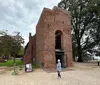 Visitors explore the ruins of a historic brick building with an adjacent large tree set against a cloudy sky