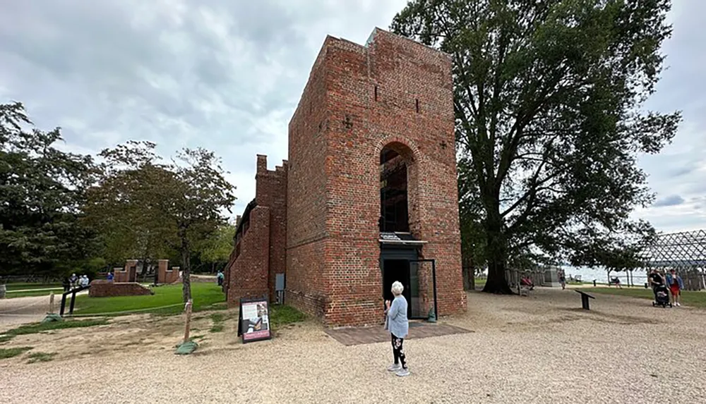 Visitors explore the ruins of a historic brick building with an adjacent large tree set against a cloudy sky