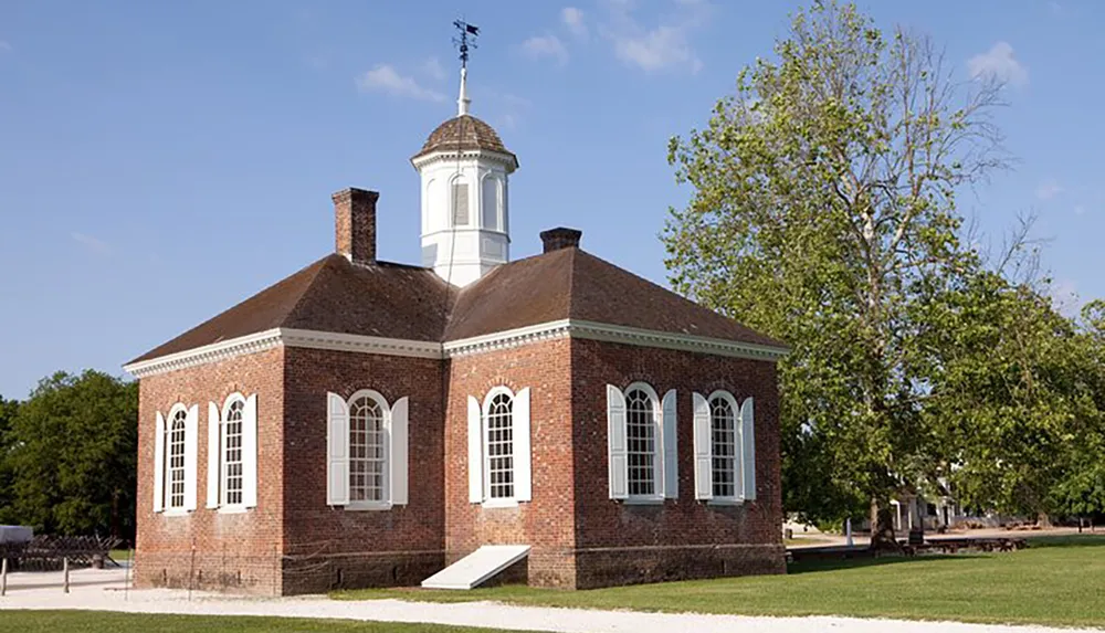 This image shows a colonial-era brick building with a white cupola set against a clear sky with greenery surrounding it