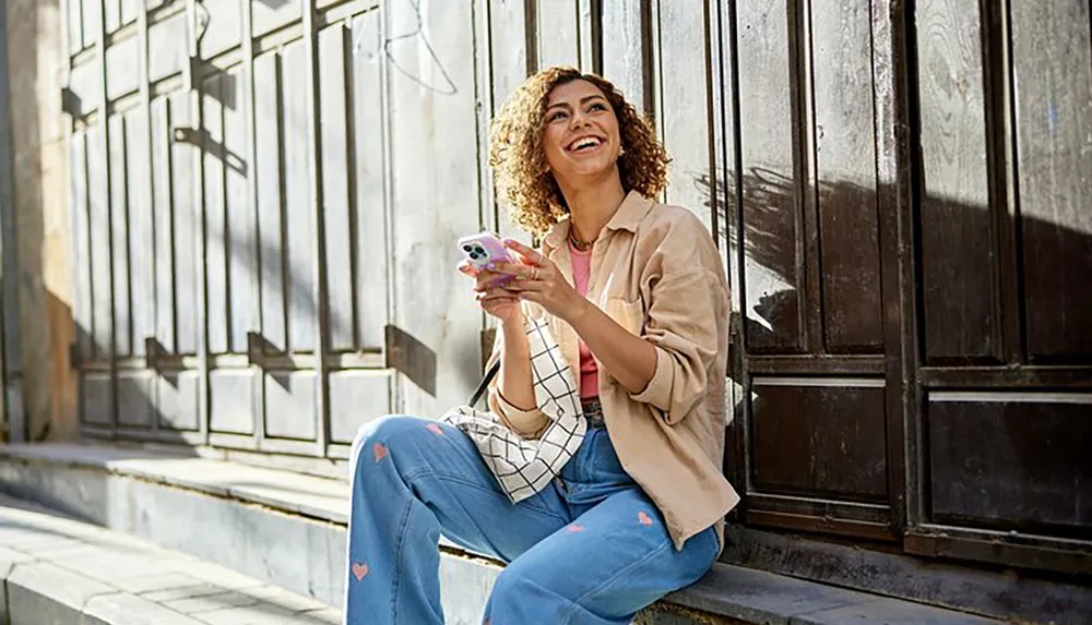 A smiling woman sits on steps outdoors holding a smartphone