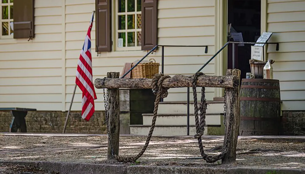 The image shows a rustic wooden well with a rope situated in front of a traditional building with an American flag conveying a sense of historical or rural American life