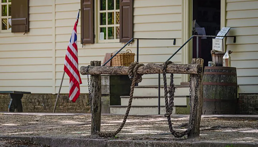 The image shows a rustic wooden well with a rope, situated in front of a traditional building with an American flag, conveying a sense of historical or rural American life.