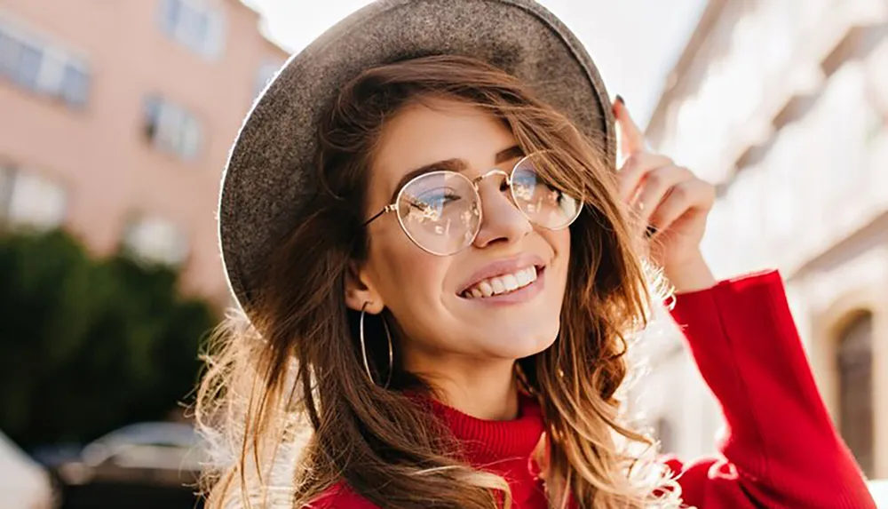 A smiling woman wearing a hat and round glasses is touching her glasses with her right hand exuding a cheerful and stylish vibe