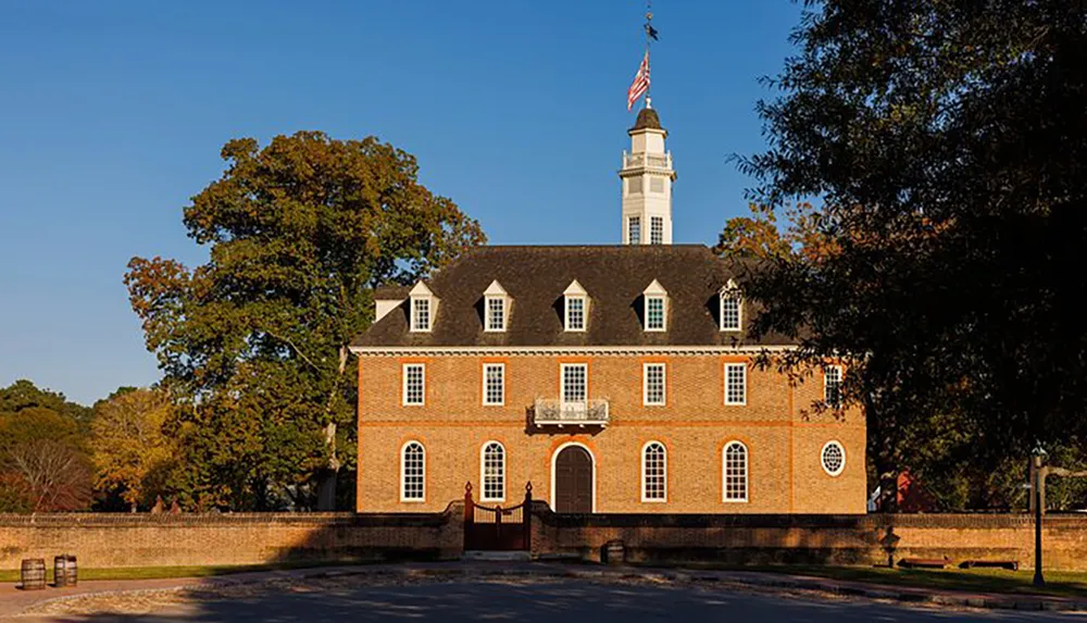 The image shows a colonial-era brick building with a central entrance flanked by windows dormer windows on the roof and a tall white steeple all set against a clear blue sky and surrounded by autumn foliage