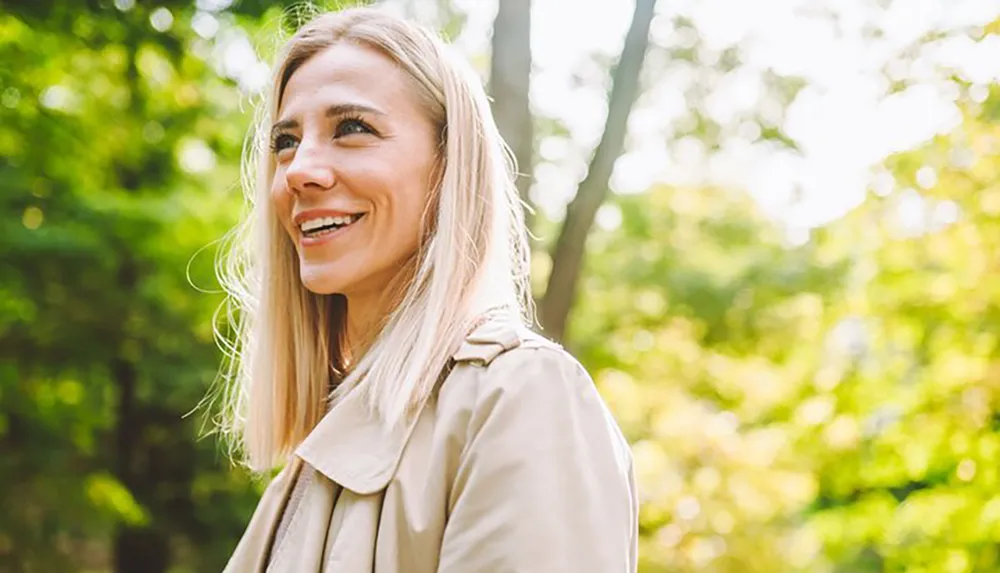 A smiling woman with blonde hair is looking away from the camera with a sunlit green forest in the background