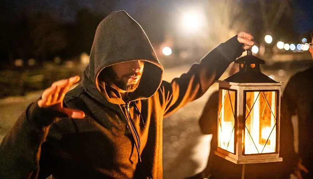 A person in a hooded jacket holds a lit lantern at night casting a warm glow in the surrounding darkness