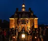 A group of people stand in front of an illuminated historic building at dusk listening to a guide dressed in period clothing