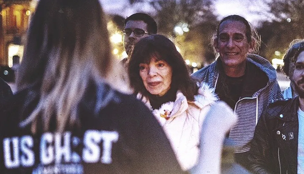 A group of people are engaged in a conversation in a dimly lit outdoor setting with one persons back to the camera featuring a jacket with the text US GHST