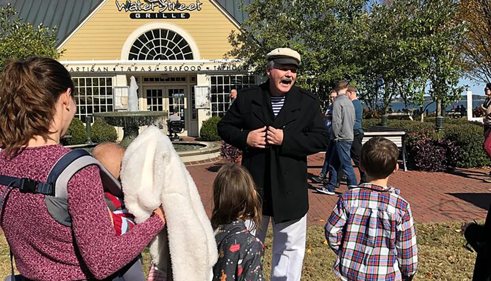 A man dressed in a black coat and beret appears to be cheerfully interacting with children in front of a restaurant named Water Street Grille on a sunny day