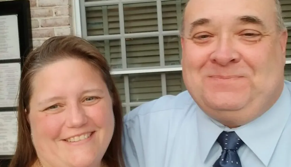 Two people are smiling for a close-up selfie with one individual wearing a blue shirt and tie and the other a darker top set against a background that appears to be the exterior of a building with shutters