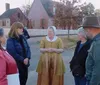 A group of people is listening to a woman dressed in historical attire possibly a reenactor or tour guide on a street that appears to come from an earlier era