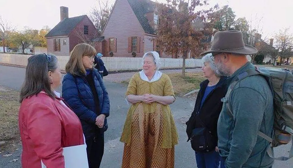 A group of people is listening to a woman dressed in historical attire possibly a reenactor or tour guide on a street that appears to come from an earlier era