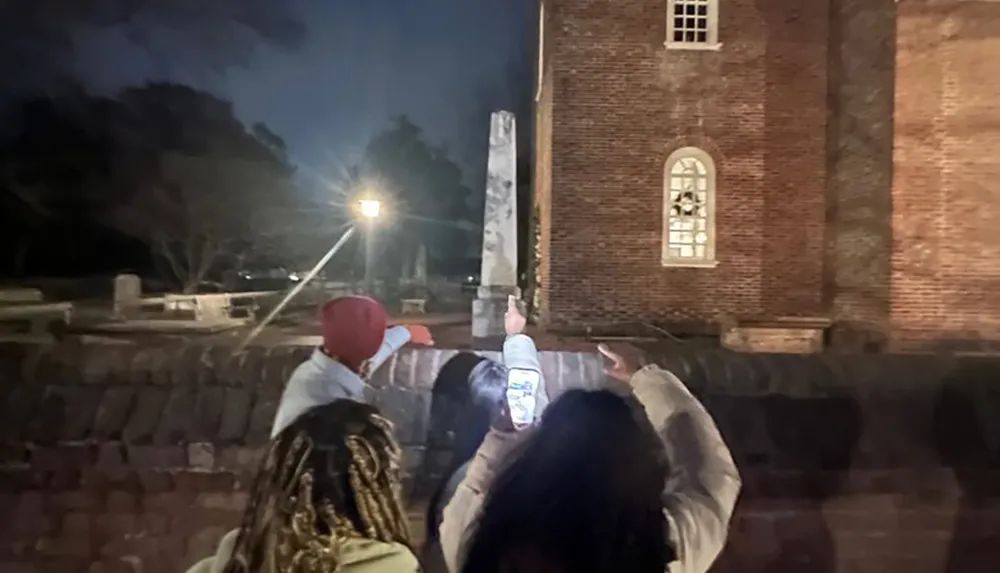 Two individuals are taking pictures of a historical building at night with one person using a smartphone while the other points a flashlight towards an obelisk or monument