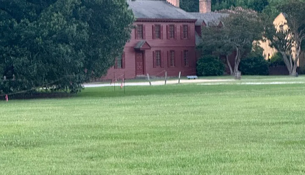 A wide lush green lawn extends towards a traditional red building with white-trimmed windows and a smaller yellow house partially visible in the background