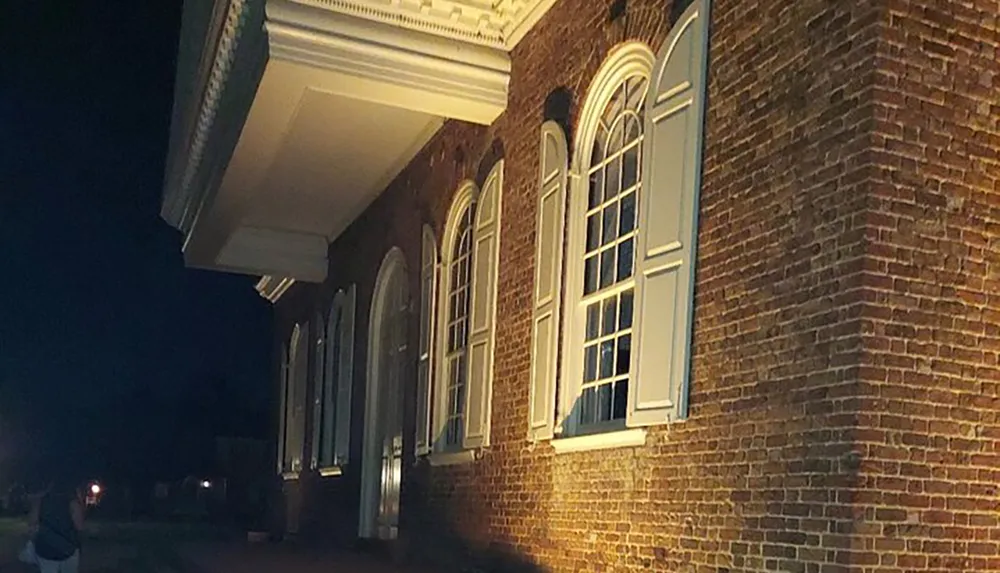 The image shows the exterior of a brick building at night with illuminated windows and a white-trimmed overhang with a persons silhouette visible in the foreground