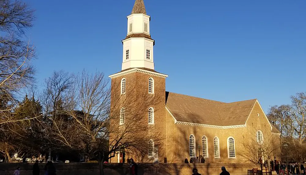 The image depicts a traditional brick church with a tall white steeple under a clear blue sky surrounded by leafless trees and a few people nearby