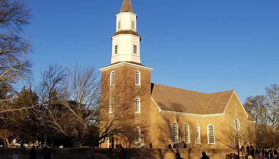 The image depicts a traditional brick church with a tall white steeple under a clear blue sky, surrounded by leafless trees and a few people nearby.