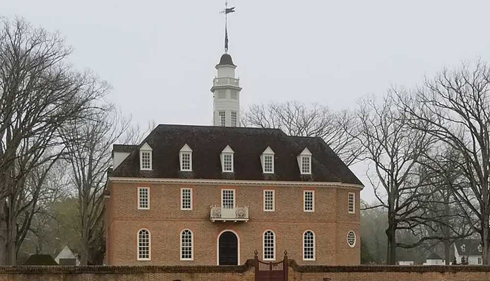 The image shows a two-story colonial brick building with a central bell tower surrounded by a wooden fence partially obscured by leafless trees under an overcast sky