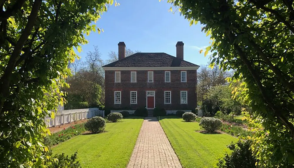 A symmetrical red-brick colonial house at the end of a brick pathway flanked by green lawns and framed by leafy trees under a clear blue sky