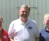 Three people are smiling and posing for a photograph in front of a white corrugated metal backdrop with two of them wearing shirts with similar circular logos