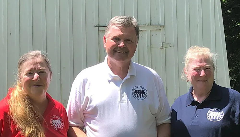 Three people are smiling and posing for a photograph in front of a white corrugated metal backdrop with two of them wearing shirts with similar circular logos