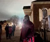 A person is smiling for the camera during a night-time outdoor event near an historic-looking building with other people and a wooden stock in the background