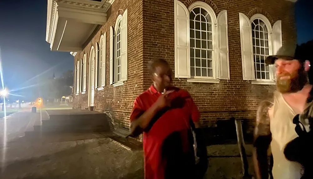 The image shows two men standing outside a brick building at night with one man gesturing mid-conversation and the other listening under artificial lighting that creates a lens flare effect