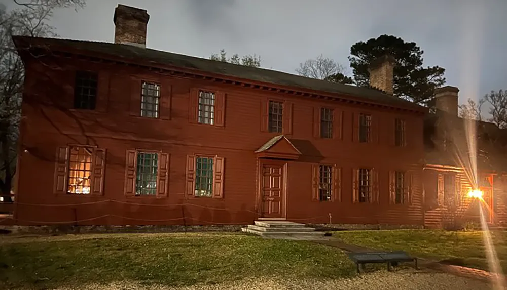 The image shows a two-story colonial-style red house with white-trimmed windows lit from the inside during dusk or night creating a warm and slightly mysterious atmosphere