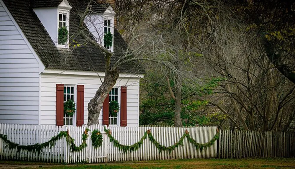 This image features a traditional white house with red shutters behind a white picket fence adorned with greenery set against a backdrop of leafless trees