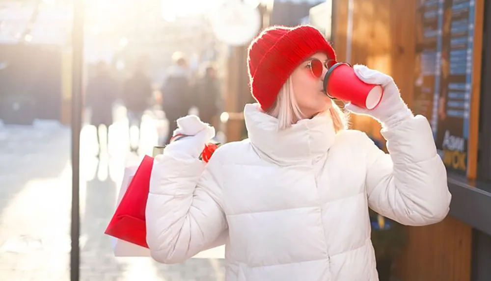 A person in a winter coat and red beanie is drinking from a red cup and carrying shopping bags on a sunlit street