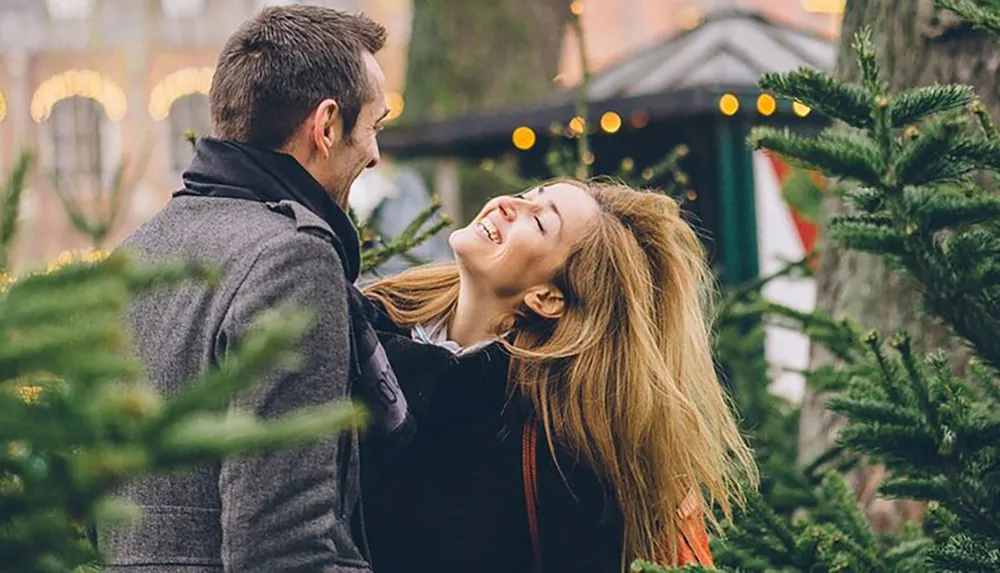 A happy couple is sharing a joyous moment together amidst a setting of evergreen trees likely representing a festive or holiday atmosphere