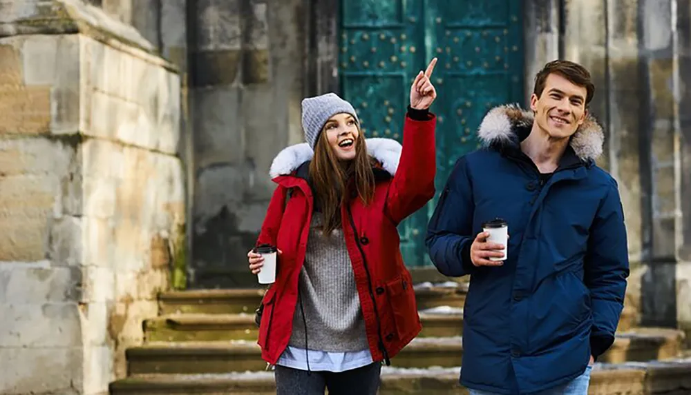 A woman pointing upwards with a smile and a man holding a coffee cup are standing in front of an old building both wearing winter jackets