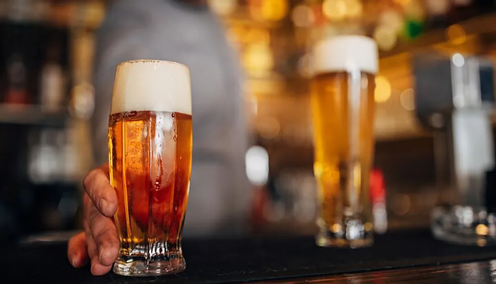 A person is holding a pint of beer with a frothy head with another full glass in the background presumably on a bar counter