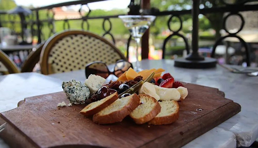 A cheese platter with a variety of cheeses fruits and sliced bread is presented on a wooden cutting board at an outdoor dining setting with a martini glass and sunglasses in the background