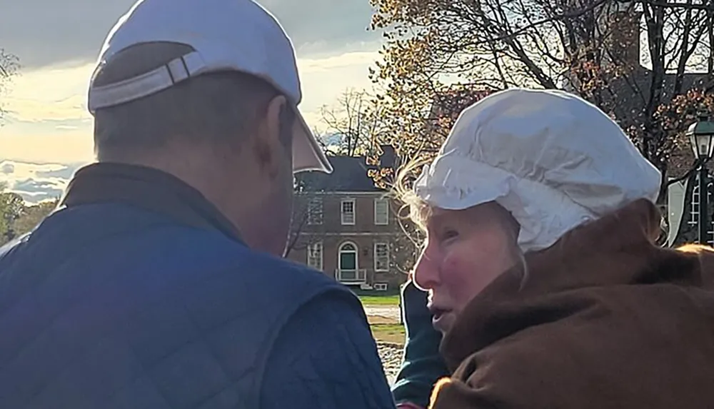 Two people are having a face-to-face conversation outdoors one is wearing a white cap that appears to be of historical styling